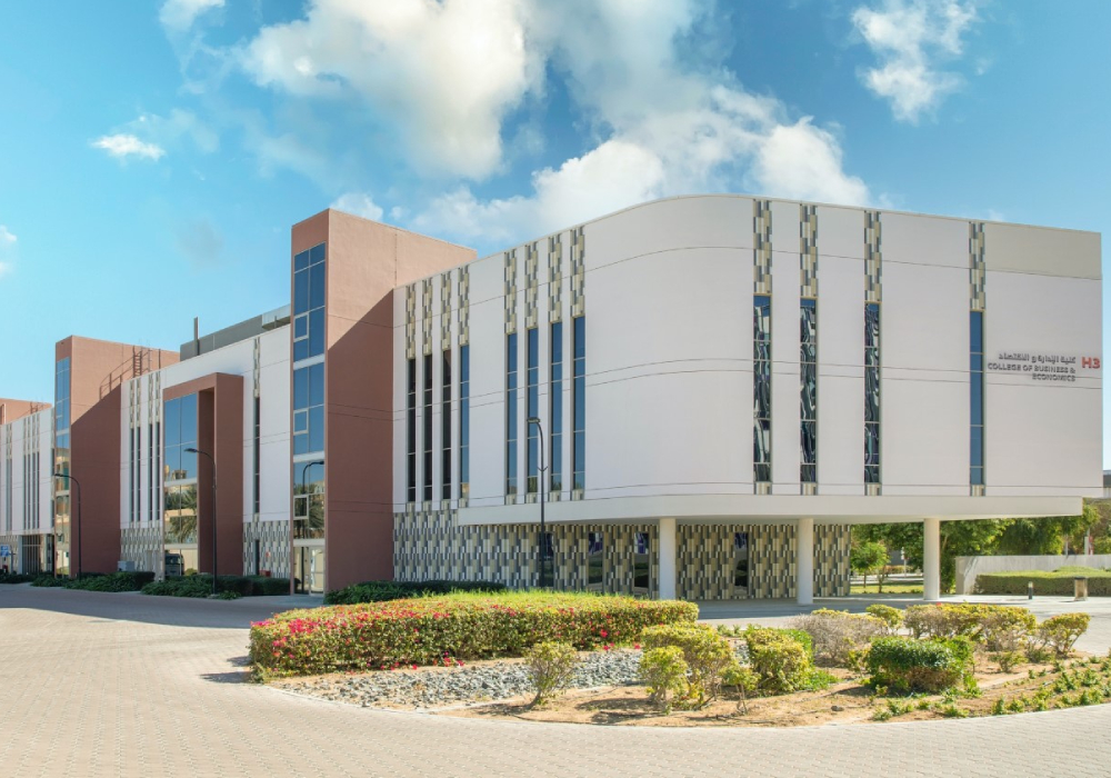 A modern academic building at the College of Business and Economics (CBE), UAEU, featuring a white and beige facade with tall vertical windows, landscaped gardens, and a clear blue sky overhead.