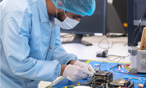 مهندس مختبر يرتدي معدات وقاية ويقوم بعناية بتركيب مكونات إلكترونية على لوحة دائرة، مما يجسد أبحاث تكنولوجيا الفضاء المتقدمة في المركز الوطني لعلوم وتكنولوجيا الفضاء