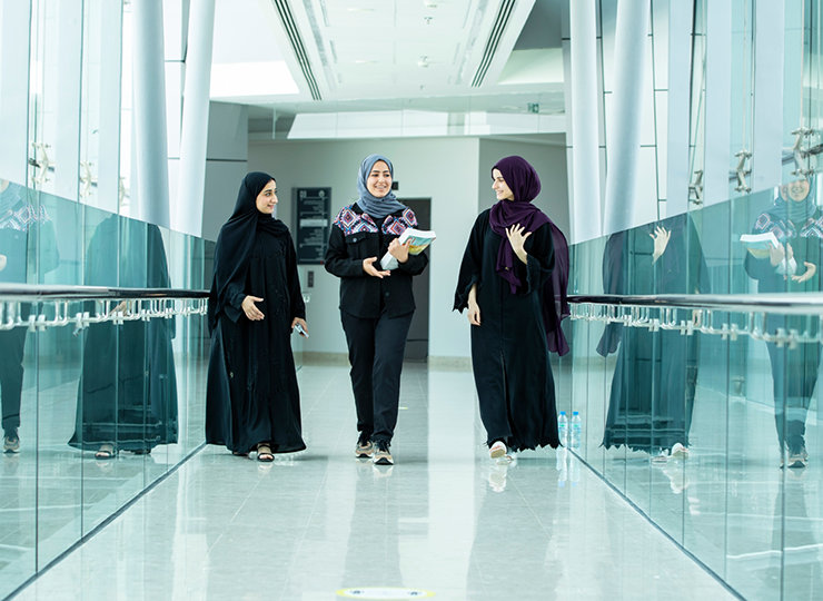 ثلاث طالبات يمشين ويتحدثن في ممر حديث مشرق ذو جدران زجاجية في جامعة الإمارات.