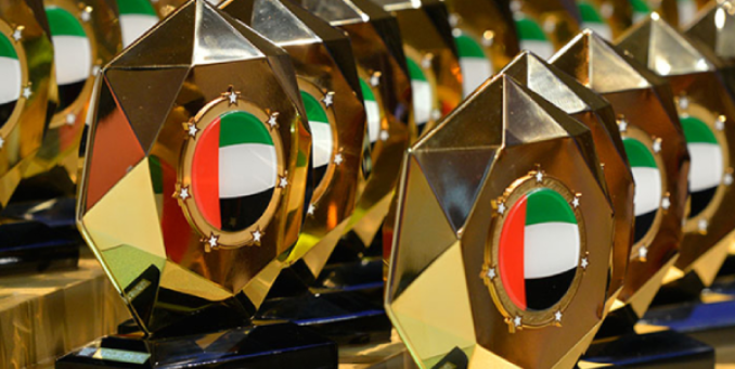 Modern campus buildings of the United Arab Emirates University with landscaped greenery and clear blue sky, featuring portraits of UAE leaders on either side of the entrance.